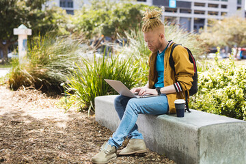 Thoughtful albino african american man with dreadlocks sitting in park using laptop