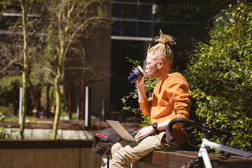 Thoughtful albino african american man with dreadlocks sitting in park drinking coffee using laptop
