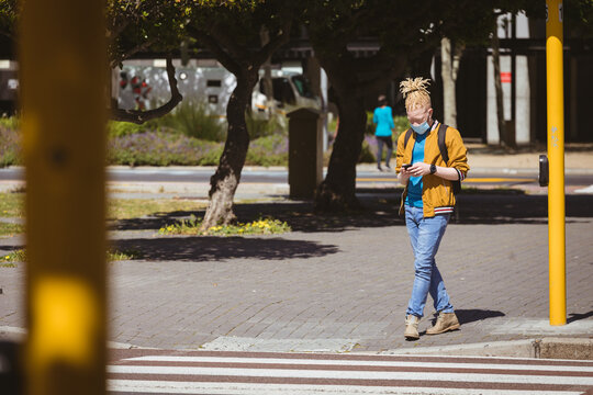 Albino african american man with face mask and dreadlocks walking and using smartphone - Powered by Adobe