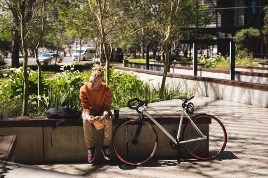 Albino african american man with dreadlocks sitting in park with bike eating sandwich