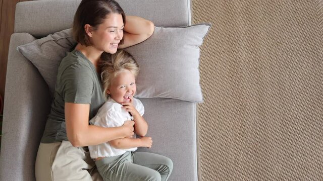Mother and daughter enjoy resting at home. spbi Smiling young housewife tickles laughing little kid girl lying on soft sofa in living room upper view