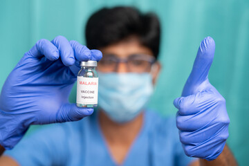 Doctor with medical face showing malaria vaccine bottle with thumbs-up gesture - concept of new...