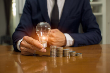 Businessman holding a bright light bulb saving bank a coins on wooden table  