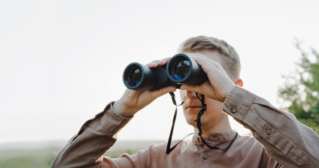 Fototapeta premium A young man looks through binoculars at a beautiful hilly landscape.The concept of hunting, travel and outdoor recreation. Banner with copy space.A traveler or hunter is observing through binoculars.