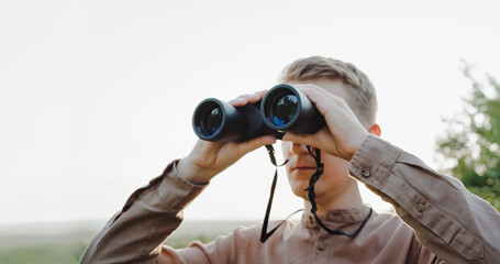 A young man looks through binoculars at a beautiful hilly landscape.The concept of hunting, travel and outdoor recreation. Banner with copy space.A traveler or hunter is observing through binoculars.