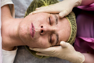 Beautician cleans the patient facial skin with massaging movements using a scrub. Top view.