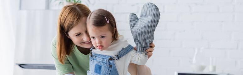 Happy mother looking at child with down syndrome and baking glove in kitchen, banner.