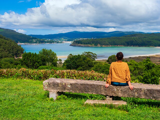 Una chica joven sentada en un banco de piedra mirando una le las rias gallegas