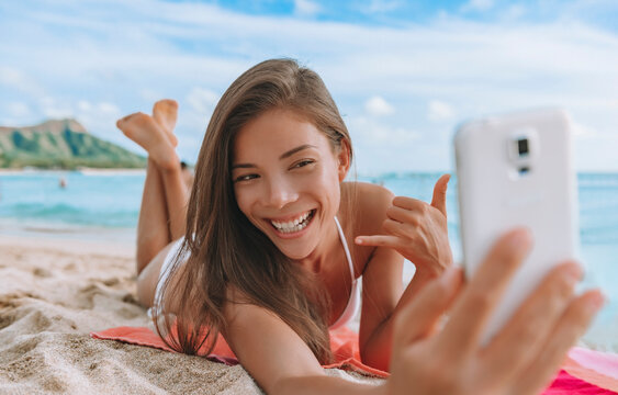 Hawaii Waikiki beach vacation Asian bikini model woman taking selfie with mobile photo lying down tanning in Honolulu. Summer travel holiday.