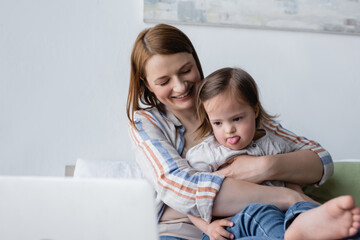 Fototapeta premium Smiling mother hugging daughter with down syndrome sticking out tongue near blurred laptop.