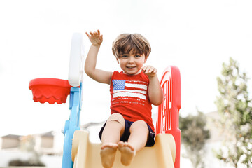 little boy playing on the backyard on a little slide with an American shirt 