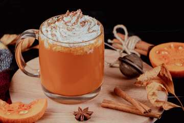 Pumpkin coffee latte with whipped cream on dark background. Hot autumn drink with cinnamon and spices in glass mug on a wooden tray.