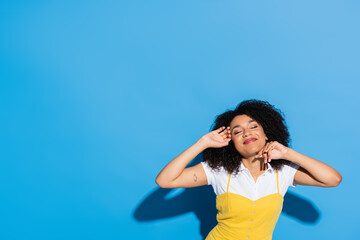 pleased african american woman posing with hands near face and closed eyes on blue