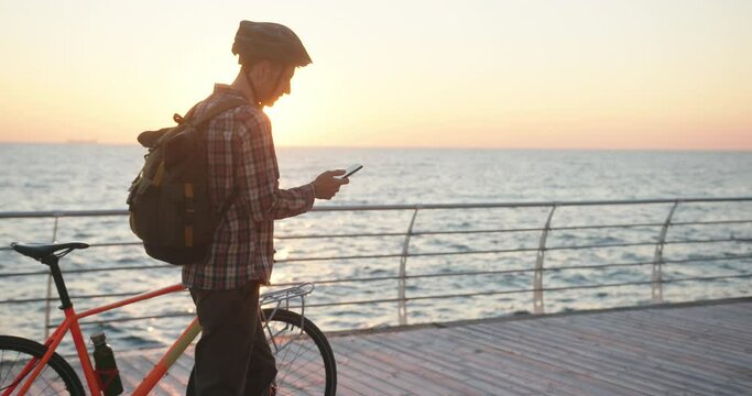 Authentic creative shot of a man with bicycle walking using his smartphone phone on sea embankment at sunrise
