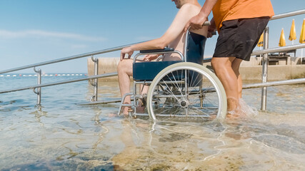 Man with disability on a wheelchair being transported into sea for swimming using a ramp.