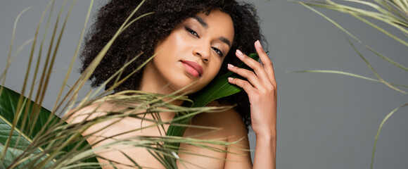 young african american woman with naked shoulders posing near green leaves isolated on grey, banner
