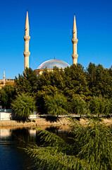 Fototapeta na wymiar Picturesquec landscape view of mosque with two minarets against blue sky. The mosque is hidden behind dense green trees. View from the bridge over Red River (Kizilirmak). Avanos, Nevsehir, Turkey