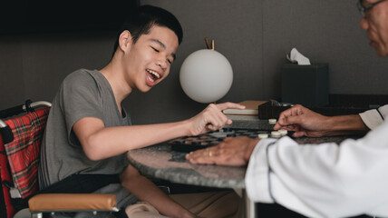A young man with disability smiling face looking and playing a checkers on a table with father in...