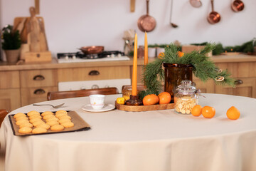Cozy festive table setting with tablecloth with xmas decorations, cookies and candles in kitchen. Concept of Christmas or New year family dinner. Branches of spruce in vase on wooden tray on table. 