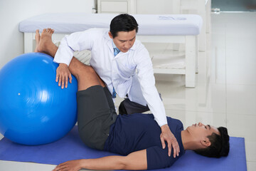 Horizontal shot of modern Asian physiotherapist helping male patient to do exercises on mat with use of ball for rehabilitation