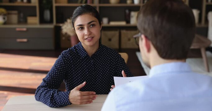 Job interview. Young indian female job seeker sit at desk in male hr office speak about herself describe experience. Confident woman candidate on vacant place answer recruiter question based on resume