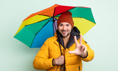 young handsome man smiling and looking friendly, showing number three. rain and umbrella concept