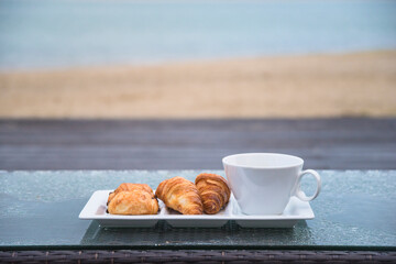 Breakfast in front of the sea; white cup of coffee and french croissants on white plate on the table in front of the sea