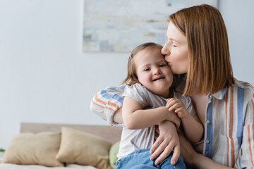 Mother kissing smiling baby daughter with down syndrome in bedroom.