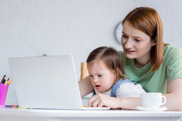 Smiling freelancer using laptop near daughter with down syndrome and coffee at home.