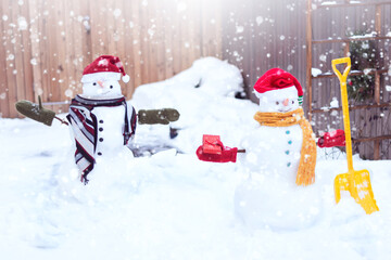 Two smiling snowmen - happy couple  in knitted mittens, scarf and red caps with gifts and shovel  in the back yard fence  background during snowfall. Winter, Christmas and New year concept  