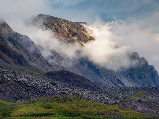 Wonderful minimalist landscape with big mountain peaks above low clouds, fog over the mountain slopes in the distance, white clouds filling the mountain gorge. Sunlight on a mountainside.