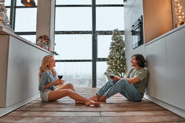 Fotobehang Young couple in love sitting on the floor, celebrating something and drinking red wine in bright kitchen. Home, love, dating, romance and christmas time concept © HBS
