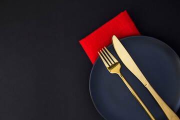 Festive table setting with gold knife and fork on the black plate on the black background. Top...
