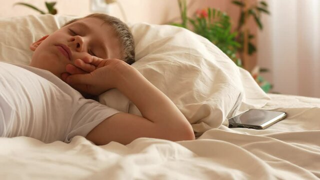 A Caucasian boy of 7-9 years old wakes up in the daylight from a smartphone alarm in his bed at home, turns off the signal and falls asleep again. the child does not want to wake up in the morning