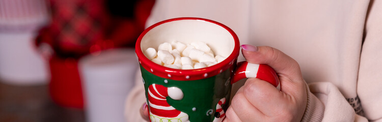 Young girl in sweater is holding a mug with hot chocolate or coffee with marshmallow. Christmas lights on, cozy holiday atmosphere, aromatherapy for cold winter season. hand with cup of hot cocoa