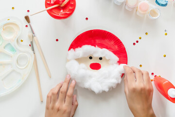 The child glues the parts Santa made from paper plate. Handmade. Project of children's creativity, handicrafts, crafts for kids. Preparation for christmas. Decoration.