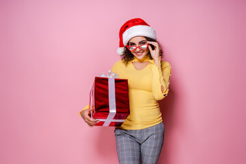 Smiling curly girl in Santa's hat, glasses holds present while stands on pink background. New Year