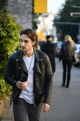Stylish man in leather jacket holing coffee to go while walking on street 