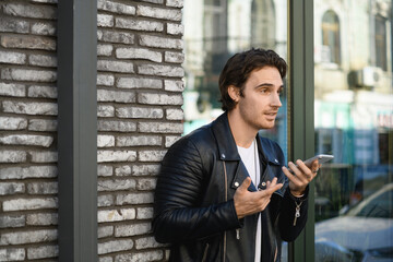 Obraz premium Good looking man in leather jacket recording voice message smartphone on street 