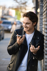 Stylish man in leather jacket recording voice message on smartphone near building 
