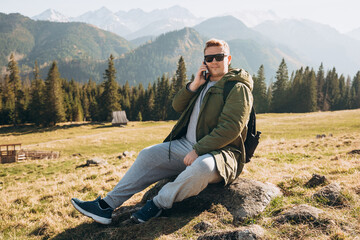 Naklejka na ściany i meble Portrait of positive man in sunglasses use smart phone over nature background. Mountain peaks. Freedom, happiness, travel and vacations concept, outdoor activities