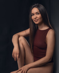 Naklejka premium Studio photo of pretty brunette woman sitting on cube and hugging her leg with hand on black background