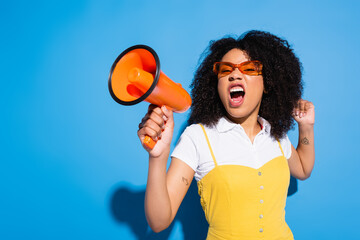 excited african american woman in eyeglasses shouting in orange loudspeaker on blue