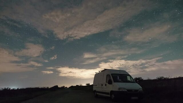 Moving clouds under stars at night sky besides a road with camper van in foreground, Brittany, France