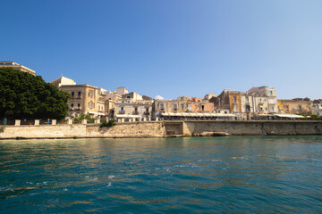 Fototapeta na wymiar Sea side view of Siracusa Ortigia island old town buildings houses and churches in sunny summer day. A bright colorful photo good for touristic booklet or book, boat trip ads, posters etc.