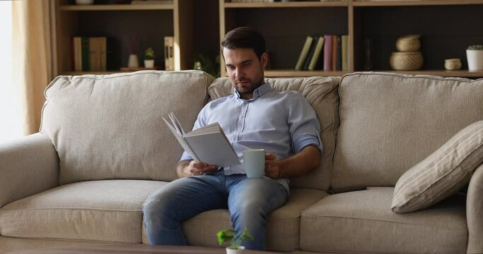 Best way to spend leisure time. Peaceful young male introvert pass free serene weekend on cozy sofa at home read favorite book drink hot tea. Calm carefree man enjoy favorite novel with cup of coffee