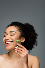 happy african american woman with perfect skin and natural makeup holding jade roller isolated on grey