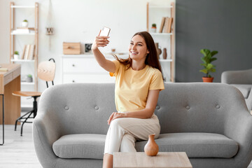 Young smiling woman sitting on sofa and taking selfie - obrazy, fototapety, plakaty