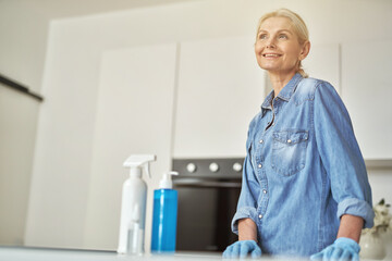 Fototapeta premium Attractive mature woman in rubber gloves smiling away, ready for clean up in the kitchen. Detergents, sanitizer spray and gel in bottle on the surface