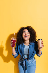 cheerful african american woman with coffee and fresh smoothie smiling at camera on yellow